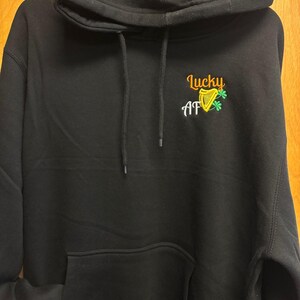 Könnte beinhalten: Ein schwarzer Kapuzenpullover mit einer Kängurutasche auf der Vorderseite und passenden schwarzen Kordelzügen. Im Brustbereich befindet sich ein gesticktes Motiv mit dem Text Lucky AF in Orange und Weiß, ergänzt durch eine gelbe Harfe und ein grünes Kleeblatt.