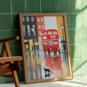Puede incluir: Una impresión enmarcada de una escena callejera londinense bajo la lluvia. Un autobús de dos pisos rojo es el elemento central, con edificios al fondo. Un gato negro está en primer plano. La obra de arte está en un marco de madera, apoyada contra una pared de azulejos verdes.