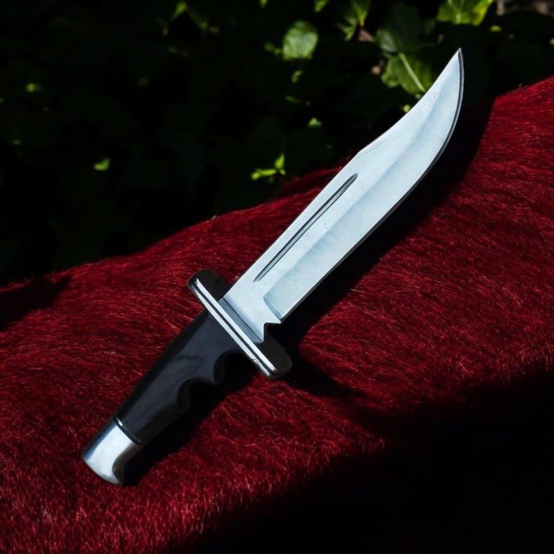 Puede incluir: Un cuchillo de caza de acero inoxidable con mango negro. La hoja es curva y afilada, reflejando la luz. El cuchillo descansa sobre una superficie roja texturizada, con un fondo verde oscuro.
