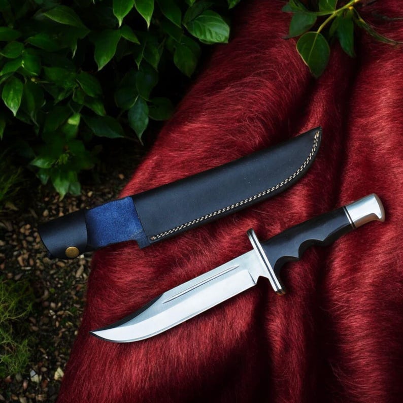 Puede incluir: Cuchillo de caza con una hoja de acero pulido y un mango negro, sobre una superficie roja. El cuchillo tiene una funda de cuero negro con detalles de ante azul. La hoja mide unos 20 cm.