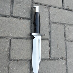 Puede incluir: Un cuchillo de caza grande con una hoja de acero pulido y un mango negro. El mango tiene ranuras para los dedos y una virola y pomo plateados. El cuchillo est&aacute; sobre una superficie de ladrillos grises.