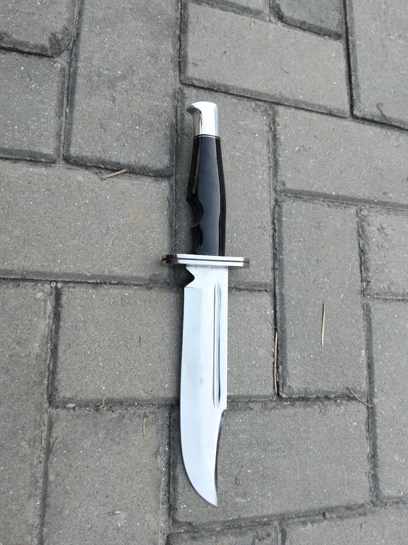 Puede incluir: Un cuchillo de caza grande con una hoja de acero pulido y un mango negro. El mango tiene ranuras para los dedos y una virola y pomo plateados. El cuchillo est&aacute; sobre una superficie de ladrillos grises.