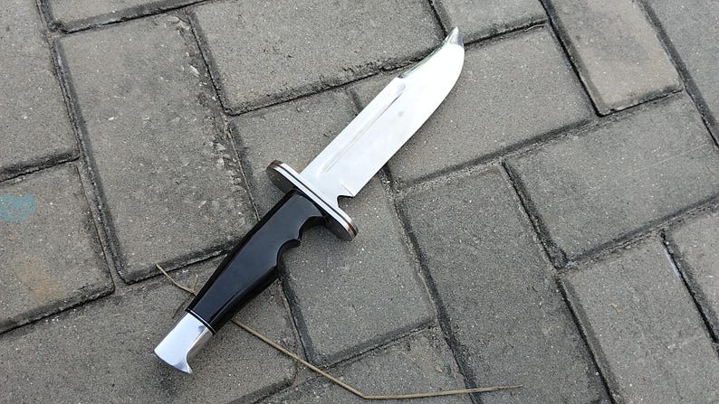 Puede incluir: Un cuchillo grande de acero inoxidable con mango negro y detalles plateados. La hoja es ancha y brillante. El mango tiene un dise&ntilde;o curvado. El cuchillo est&aacute; sobre una superficie de ladrillos grises.