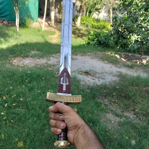 May include: A silver and gold sword with a trident symbol on the hilt. The sword has a long, pointed blade and a detailed handle. The handle is gold with the word "ANAKAYMOS" engraved on it. The sword is held in a hand against a green background.