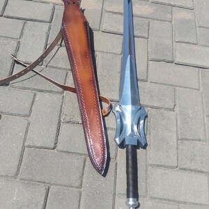 Puede incluir: Una espada plateada con empu&ntilde;adura negra y funda de cuero marr&oacute;n. La espada tiene una hoja ancha y plana y una guarda decorativa. La funda tiene una correa para llevarla.