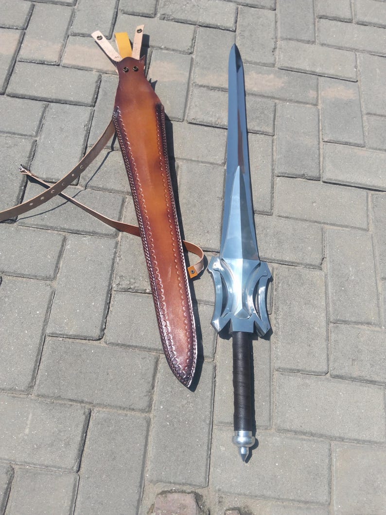 Puede incluir: Una espada plateada con empu&ntilde;adura negra y funda de cuero marr&oacute;n. La espada tiene una hoja ancha y plana y una guarda decorativa. La funda tiene una correa para llevarla.