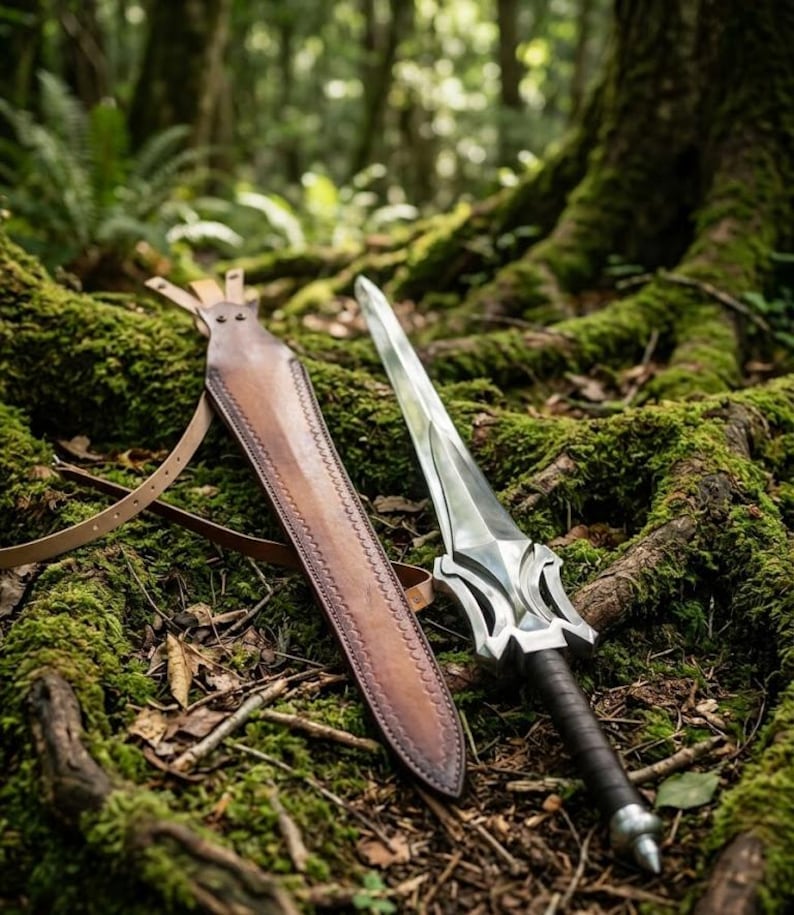 Puede incluir: Una espada plateada y negra con una funda de cuero descansa sobre un suelo forestal cubierto de musgo. La espada tiene una empu&ntilde;adura detallada y una hoja afilada. La funda de cuero marr&oacute;n est&aacute; abierta, revelando toda la longitud de la espada.