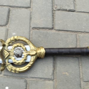 Puede incluir: Una espada decorativa con una empuñadura dorada y un mango marrón oscuro. La empuñadura presenta un diseño ornamentado con una piedra central. La hoja es plateada. La espada está sobre una superficie de ladrillos grises.