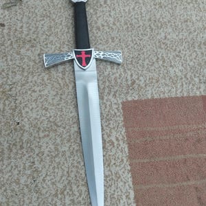 May include: A decorative sword with a silver blade and guard, featuring a red cross. The handle is black, and the pommel has a red jewel. The sword is a historical replica.