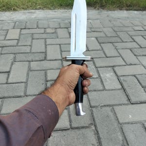 Puede incluir: Un cuchillo grande de acero inoxidable con una hoja curva y un mango negro. El cuchillo tiene una guarda plateada y un dise&ntilde;o decorativo en la hoja. El mango se sostiene en una mano.