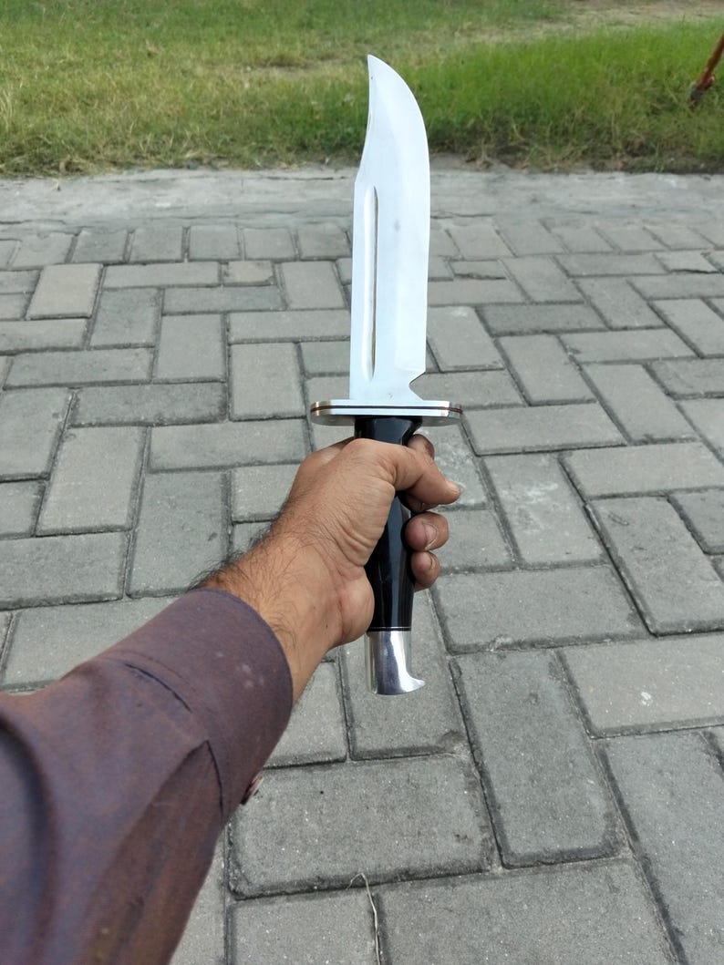 Puede incluir: Un cuchillo grande de acero inoxidable con una hoja curva y un mango negro. El cuchillo tiene una guarda plateada y un dise&ntilde;o decorativo en la hoja. El mango se sostiene en una mano.