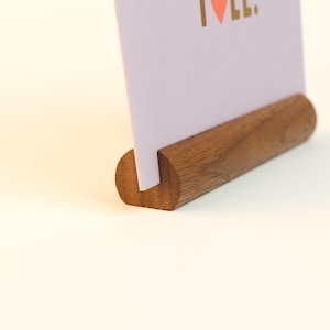 Könnte beinhalten: Ein Kartenhalter aus Holz mit einer hellvioletten Karte. Die Karte hat goldenen Text und ein kleines rosa Herz. Der Holzhalter ist dunkelbraun und hat eine abgerundete Form.