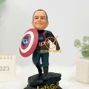 May include: A Captain America bobblehead figurine with a realistic head and a shield featuring the "Five9" logo. The figurine is on a black base with the words "Let's Go!" in gold lettering. The figurine is wearing a blue and red costume.