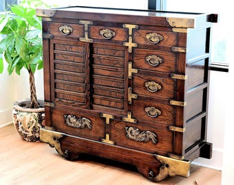 May include: A dark brown wooden cabinet with brass hardware, including handles and corner accents. The cabinet features multiple drawers and a sliding door with horizontal slats. A decorative plant in a patterned pot is to the left.