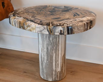 May include: A unique side table with a round top made from petrified wood, showcasing a natural pattern of browns, grays, and creams. The table is supported by a cylindrical base with a marbled appearance. The table's dimensions are approximately 24 inches in height.