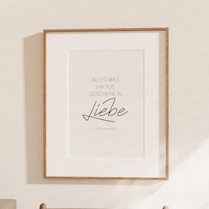 Könnte beinhalten: Ein gerahmter Druck mit den Worten "ALLES WAS IHR TUT, GESCHEHE IN Liebe" in schwarzer Schrift. Der Druck befindet sich in einem weißen Passepartout mit einem hellbraunen Holzrahmen. Der Druck hängt an einer weißen Wand.