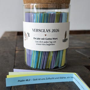 Pode incluir: Um frasco de vidro com tampa de cortiça cheio de papéis coloridos. O frasco tem um rótulo com o texto "VERSGLAS 2026" e texto em alemão. Alguns papéis são visíveis fora do frasco, com texto do Salmo 46:2. O frasco está sobre uma superfície de madeira.