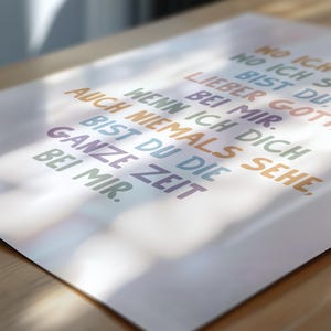 May include: A white poster with colorful text in German. The text includes phrases like "Lieber Gott bei mir" and "Ganze Zeit bei mir." The words are in various colors, including blue, orange, and green. The poster is on a wooden surface.