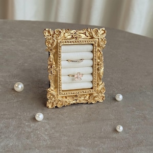 May include: A gold-colored, ornate picture frame repurposed as a ring holder. The frame holds white ring cushions, displaying two rings: one with small stones and a pink flower ring. Several white pearls are scattered around the frame.