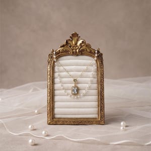 May include: A gold-colored, ornate jewelry display frame holds two necklaces. The top necklace is gold with a rectangular, clear stone pendant. The bottom necklace is a string of pearls. The frame sits on a white surface, with pearls scattered around.