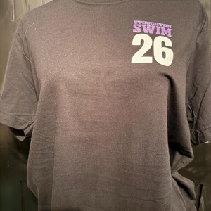 Peut inclure: Un t-shirt noir avec l'inscription "STOUGHTON SWIM 26" imprim&eacute;e en blanc et violet. Le t-shirt est pr&eacute;sent&eacute; sur un mannequin noir.