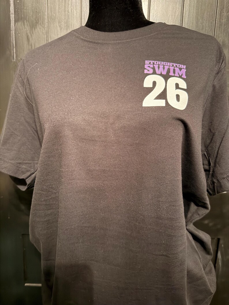 Peut inclure: Un t-shirt noir avec l'inscription "STOUGHTON SWIM 26" imprim&eacute;e en blanc et violet. Le t-shirt est pr&eacute;sent&eacute; sur un mannequin noir.