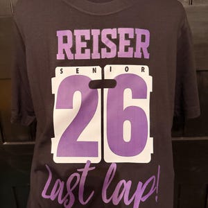 Peut inclure: T-shirt noir avec le nom "REISER SENIOR 26" en lettres violettes et blanches. Les mots "last lap!" sont &eacute;crits en violet en dessous du chiffre 26. Le t-shirt est pr&eacute;sent&eacute; sur un mannequin noir.