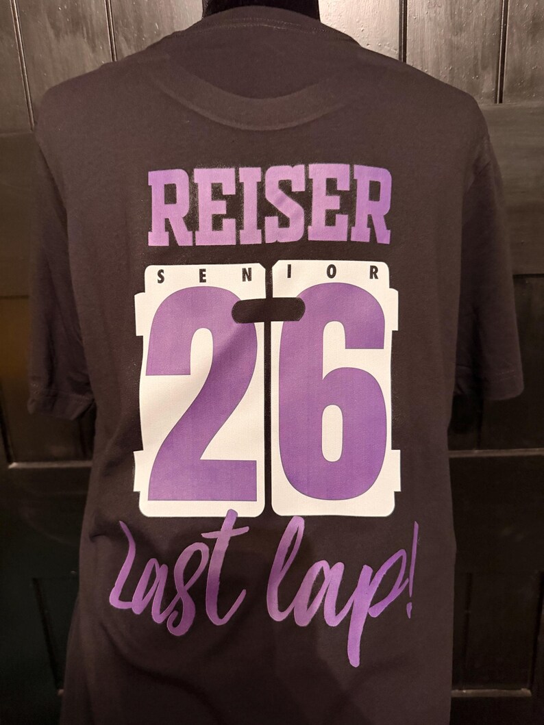 Peut inclure: T-shirt noir avec le nom "REISER SENIOR 26" en lettres violettes et blanches. Les mots "last lap!" sont &eacute;crits en violet en dessous du chiffre 26. Le t-shirt est pr&eacute;sent&eacute; sur un mannequin noir.