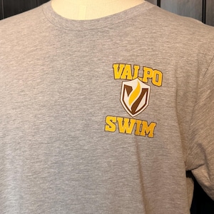 Peut inclure: Un t-shirt gris chin&eacute; avec l'inscription "VALPO SWIM" en jaune et marron. Le texte est imprim&eacute; sur le devant du t-shirt, avec un motif en forme de bouclier. Le t-shirt est pr&eacute;sent&eacute; sur un mannequin.