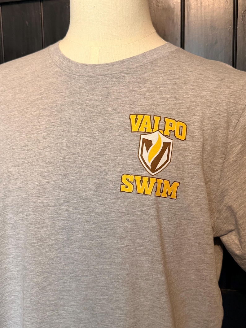 Peut inclure: Un t-shirt gris chin&eacute; avec l'inscription "VALPO SWIM" en jaune et marron. Le texte est imprim&eacute; sur le devant du t-shirt, avec un motif en forme de bouclier. Le t-shirt est pr&eacute;sent&eacute; sur un mannequin.