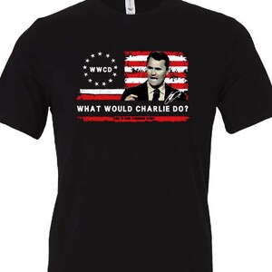 May include: Black t-shirt featuring a graphic of a man in a suit with a flag design. The flag design includes stars and stripes, with the text "WWCD" and "WHAT WOULD CHARLIE DO?" printed on it.