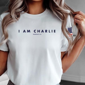 Könnte beinhalten: Weißes T-Shirt mit dem marineblauen Text "I AM CHARLIE" und "Philippians 3:14" darunter. Das Shirt hat kurze Ärmel und einen Rundhalsausschnitt. Auf dem Ärmel ist eine amerikanische Flagge abgebildet.
