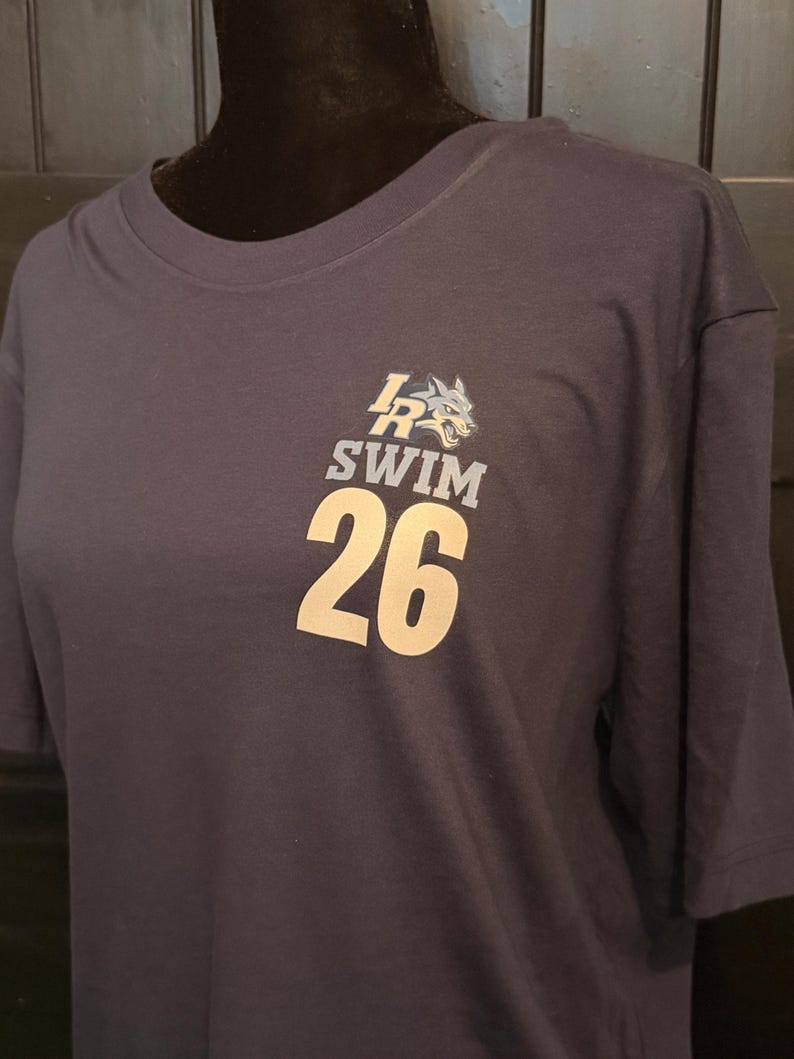 Peut inclure: T-shirt bleu marine avec un motif de t&ecirc;te de loup et le texte "LR SWIM 26" en blanc. Le t-shirt est pr&eacute;sent&eacute; sur un mannequin. Le t-shirt est fait d'une mati&egrave;re douce.