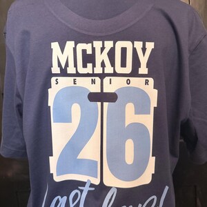 Peut inclure: Un t-shirt bleu marine avec le nom "MCKOY" en lettres blanches, le mot "SENIOR" au-dessus d'un grand chiffre "26" en bleu clair et blanc, et la phrase "Last lap!" en &eacute;criture cursive.