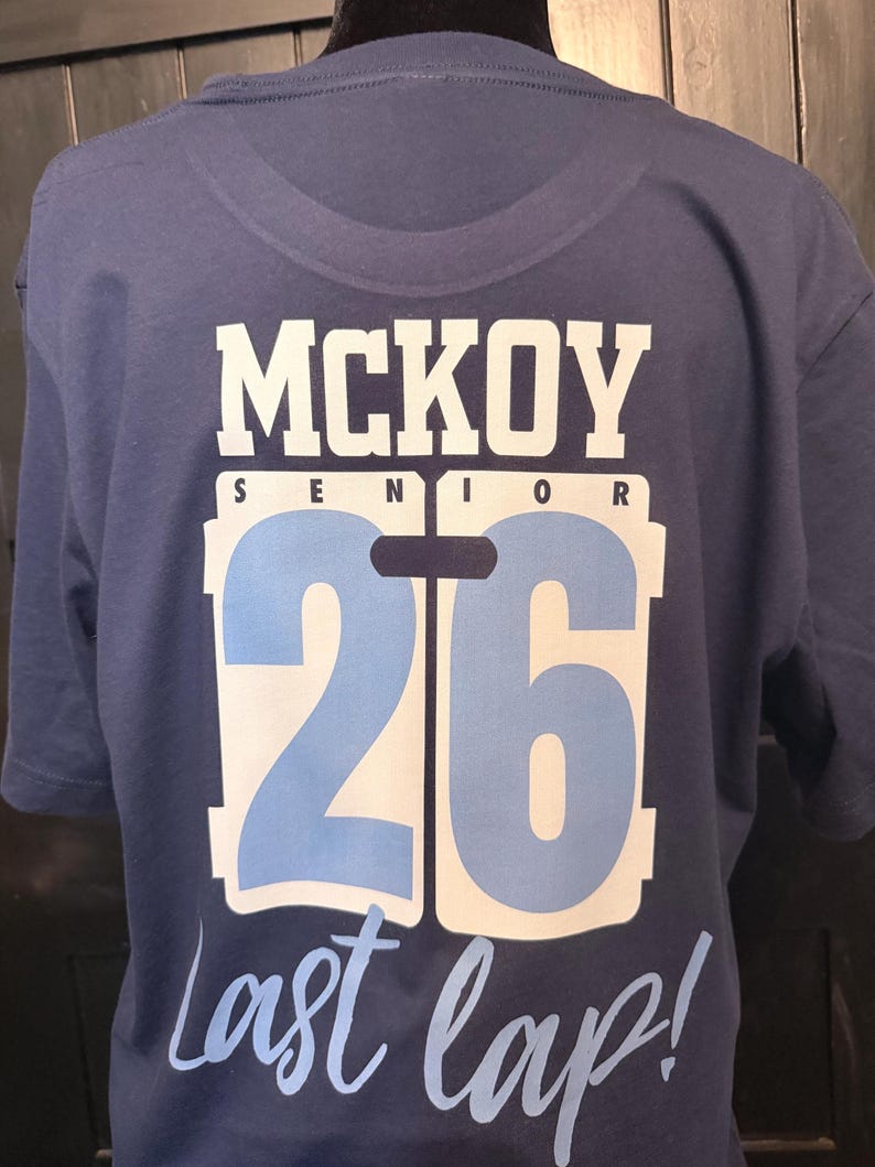 Peut inclure: Un t-shirt bleu marine avec le nom "MCKOY" en lettres blanches, le mot "SENIOR" au-dessus d'un grand chiffre "26" en bleu clair et blanc, et la phrase "Last lap!" en &eacute;criture cursive.