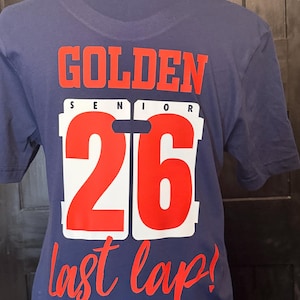 Peut inclure: T-shirt bleu marine avec les mots "GOLDEN SENIOR 26 last lap!" en rouge et blanc. Le texte est imprim&eacute; au dos du t-shirt. Le t-shirt est pr&eacute;sent&eacute; sur un mannequin.