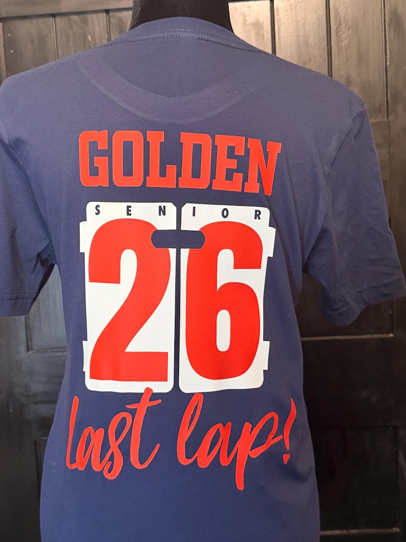 Peut inclure: T-shirt bleu marine avec les mots "GOLDEN SENIOR 26 last lap!" en rouge et blanc. Le texte est imprim&eacute; au dos du t-shirt. Le t-shirt est pr&eacute;sent&eacute; sur un mannequin.