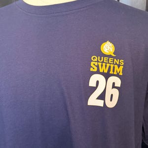 Peut inclure: T-shirt bleu marine avec l'inscription "QUEENS SWIM 26" en jaune et blanc. Le motif comprend un embl&egrave;me jaune en forme de couronne. Le t-shirt est pr&eacute;sent&eacute; sur un mannequin.