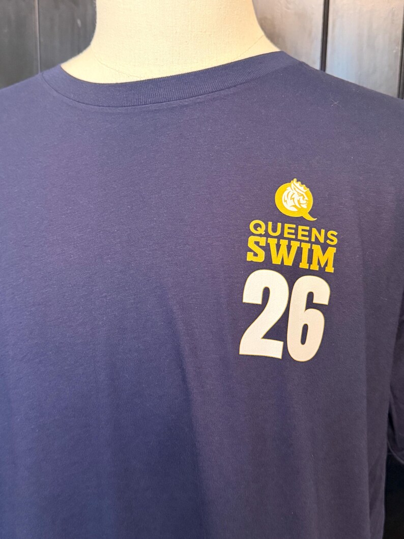 Peut inclure: T-shirt bleu marine avec l'inscription "QUEENS SWIM 26" en jaune et blanc. Le motif comprend un embl&egrave;me jaune en forme de couronne. Le t-shirt est pr&eacute;sent&eacute; sur un mannequin.