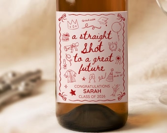 May include: A wine bottle with a gold cap and a label that reads "a straight shot to a great future." The label is red with white text and illustrations. A clear wine glass sits next to the bottle. The text "Congratulations Sarah Class of 2026" is also on the label.
