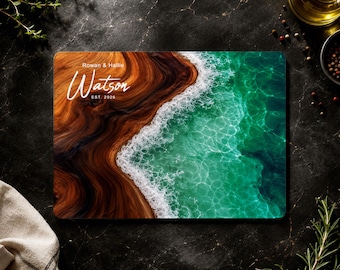 May include: A rectangular cutting board featuring a printed design of a dark wood shoreline meeting turquoise ocean waves with white sea foam. The text "Rowan & Hallie", "Watson", and "EST. 2026" is printed in white on the wood section.