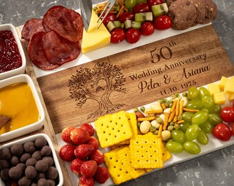 May include: A wooden charcuterie board with a white marble stripe, engraved with "50th Wedding Anniversary Peter & Ariana 11.10.1975". The board is filled with cheese, crackers, fruit, salami, and chocolate-covered snacks.