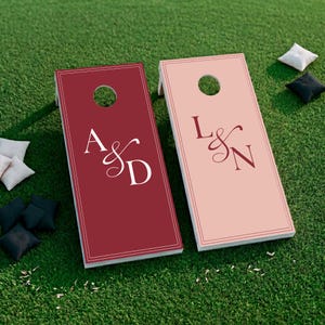 May include: Two rectangular cornhole boards with circular holes at the top. The left board is dark red with white text reading A & D. The right board is light pink with dark red text reading L & N. Both boards feature a thin rectangular border.