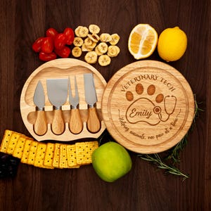 May include: A wooden cheese board set with four cheese knives and a round cutting board. The round board is engraved with the words "Veterinary Tech" and a paw print design. Fresh fruit and crackers are arranged around the boards.