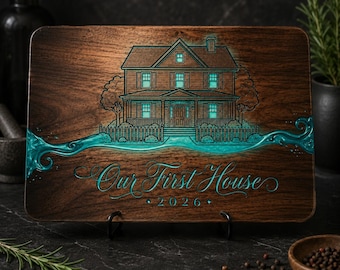 May include: A rectangular dark wood cutting board featuring an engraved illustration of a two-story house with glowing teal windows. A flowing teal resin inlay runs across the center. The text Our First House 2026 is engraved in teal script.