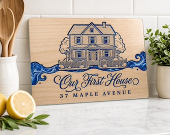 May include: A rectangular light-colored wooden cutting board featuring a blue engraved illustration of a two-story house with a picket fence. Below the house, the text reads "Our First House" in script and "37 MAPLE AVENUE" in block letters.