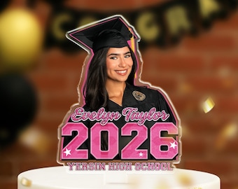 May include: A graduation cake topper featuring a smiling woman in a graduation cap and gown. The topper includes the name "Evelyn Taylor" and the year "2026" in pink, along with "YERSIN HIGH SCHOOL".