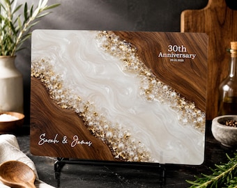 May include: A rectangular wooden serving board featuring a central white resin inlay with gold leaf flakes and embedded white pearls. The board is printed with the text "30th Anniversary 09.25.2026" and the names "Sarah & James" in white script.