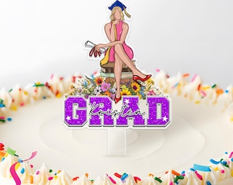 May include: A cake topper featuring a cartoon illustration of a graduate in a pink dress and cap, sitting on books and flowers. The topper has the word "GRAD" in purple glitter with the name "Kristen" in script.