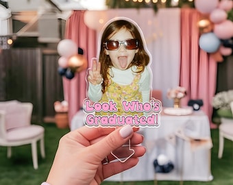 May include: A photo prop featuring a young girl wearing sunglasses and sticking out her tongue. The prop has the text "Look Who's Graduated!" in pink and white. The background includes pink curtains and balloons.